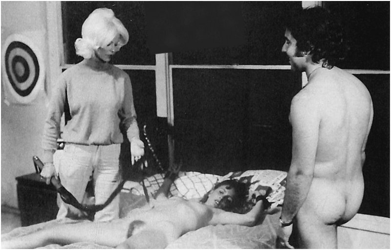 naked man and a fully clothed woman stand over a woman in bondage who is about to get a flogging